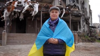 Omzunda Ukrayna bayrağı olan yaşlı ve yorgun bir kadın yıkılmış sivil evin arka planında kameraya bakıyor. Yakınlaştır ve bombalanmış harabe şehirde poz veren depresif emekliliğe odaklan.