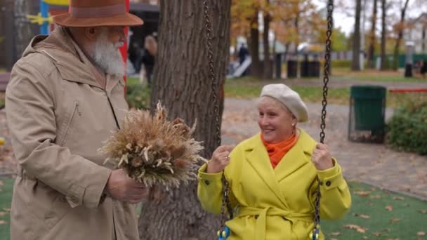 长胡子的老人站在操场上 手里拿着一束麦束 就像无忧无虑的女人荡秋千 快乐轻松的白种人老夫妇在秋天的公园里享受户外约会的快乐 — 图库视频影像