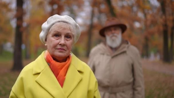 漂亮的老妇人站在室外看着相机 背后有模糊的男人 秋日公园里与难以辨认的配偶慢镜头的自信的白人退休人员的画像 — 图库视频影像