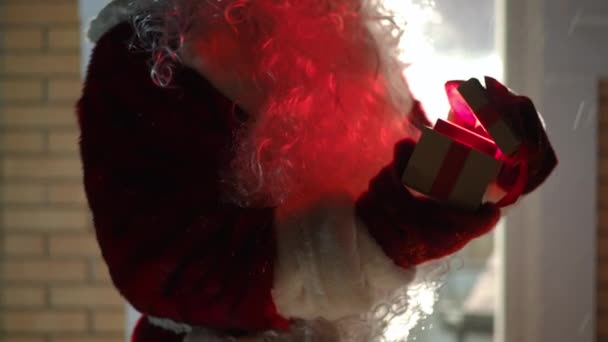 无法辨认的圣诞老人打开礼品盒 礼品盒里装着亮光 把礼物伸向相机 圣诞之夜 身穿红色服装 头戴手套的男人站在外面的雪地后院里 表现出惊讶 — 图库视频影像