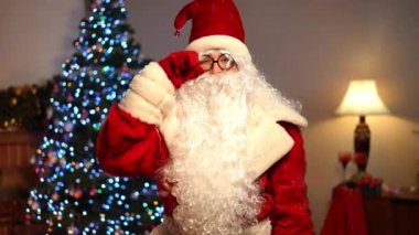 Noel Baba kostümü giymiş beyaz sakallı, gözlüklü beyaz bir adam oturma odasından çıkarken kameraya bakıyor. Noel arifesinde evde poz veren kendine güvenen genç bir adamın portresi.