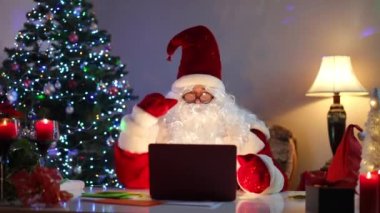 Noel Baba bilgisayarında görüntülü konuşma yapıyor. Oturma odasında oturmuş konuşuyor. Tatil arifesinde internetten kostüm giymiş beyaz adamın portresi.