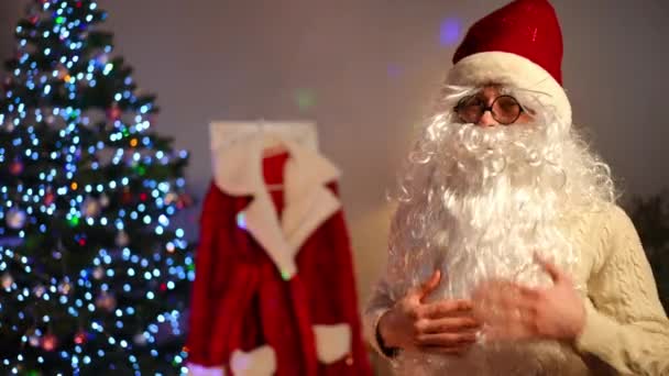 身穿白色胡须眼镜 头戴新年帽 面带微笑的男人回头望着圣诞老人的外套 转身望着镜头中的手势 圣诞期间 积极自信的年轻人在室内摆姿势的画像 — 图库视频影像