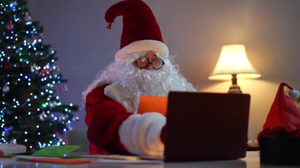 サンタクロースは手紙を読んで注文すると 屋内のテーブルでラップトップに座ってオンラインプレゼントを提示します 赤い衣装を着た白人男性の肖像画が願いをかなえる 休日と奇跡の概念 — ストック動画