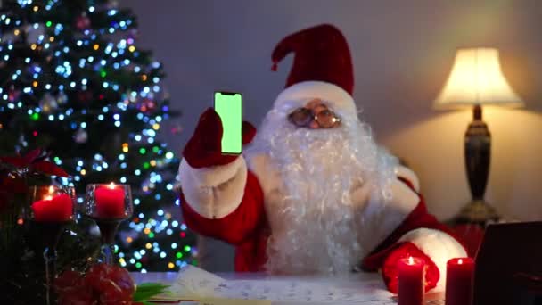 新年の木のあるリビングルームに屋内に座っているクロマキースマートフォンを示すぼやけたサンタ 赤いスーツの広告のWebテンプレートで肯定的な自信を持った男 クリスマスと現代の技術 — ストック動画