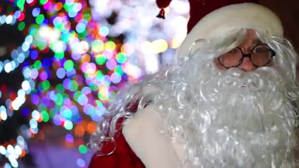 サンタの衣装を着た陽気な男が背景にクリスマスの光と夜の街の通りに立って笑顔を振っています 正の喜びの男楽しむ新年の夜屋外 — ストック動画