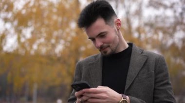 Mutlu, genç, yakışıklı, beyaz adam gülen akıllı telefon uygulamasında sonbahar ormanı mesajlaşması yapıyor. Kendine güvenen esmer adamın portresi parktaki bulutlu havanın keyfini çıkarıyor sosyal medya kayıyor.