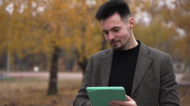 Dijital tablette internette sörf yapan gülümseyen genç adamın portresi sonbahar ormanında duruyor. Parkta internetten kendine güvenen beyaz bir adam mesajlaşırken mutlu ol. Rahatlık ve yaşam tarzı