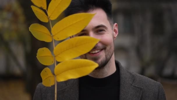 前景色 英俊的男子面带微笑 黄色的落树叶望着相机 在阴天 快乐而自信的白人小伙子在秋天的公园里摆姿势 — 图库视频影像