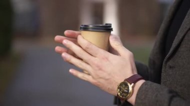 Yakın çekimde, erkek elleri açık havada tek kullanımlık kahve fincanı taşıyor. Bulutlu bir sonbahar gününde sıcak içecekle caddede duran tanınmamış beyaz genç adam.