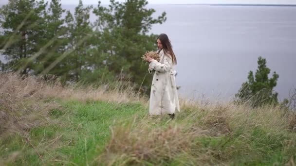 年轻的女人带着麦捆走在秋天田边的河边 环顾四周 快乐而轻松的白人女士在户外的草地上享受着宁静 慢动作 — 图库视频影像