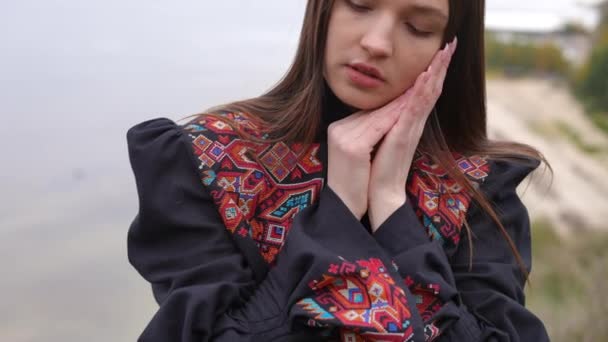 身穿绣花衣服 心事重重的年轻女子站在河岸上 慢吞吞地环顾四周 乌克兰秋日 一位身穿民族服装的美丽的黑发女士的画像 — 图库视频影像
