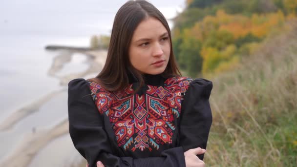 刺繍された黒いドレスの若い女性が離れて考えを見て 遅い動きで笑みを浮かべてカメラを見て ズームで信頼できる魅力的なウクライナ人女性のポートレート上の川の銀行 — ストック動画