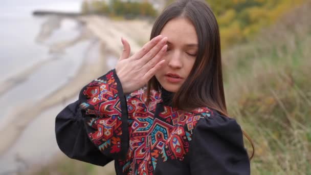 魅力的な若いブルネットの女性のズームは離れて見てスローモーションで手を移動します 美しいウクライナ人女性で国民の刺繍ドレス上の秋の曇りの日に川の銀行 — ストック動画