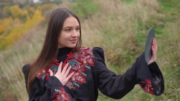 手鏡で反射を賞賛真珠のネックレスに触れるウクライナの伝統的な刺繍のドレスで美しい女性を笑顔 秋の日に野外で自信を持って魅力的な女性の肖像画 — ストック動画