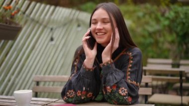 Dışarıda oturmuş telefonda konuşan dişlek gülüşlü çekici genç bir kadın. Ulusal Ukrayna nakışlı tişörtlü mutlu güzel bir kadının portresi akıllı telefonuyla sohbet ediyor, gülümsüyor.