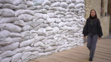 Geniş açılı, kendine güvenen, nakışlı tişörtlü genç bir kadın Ukrayna şehrindeki koruyucu kum torbalarından oluşan duvardan ağır çekimde ayrılıyor. Sokakta yürüyen güzel bayan etrafa bakıyor.