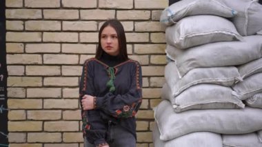 Ukrayna şehrinde kum torbalarıyla duvarda dikilmiş nakışlı gömlekli güzel bir kadının portresi. Kahverengi gözlü, kendine güvenen esmer kadının orta boy görüntüsü.