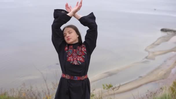 ゆったりと幸せなウクライナの若い女性の肖像刺繍黒のドレススローモーションで目を閉じて手を上げる 秋の屋外で川の背景にある無料の自信のある女性 — ストック動画