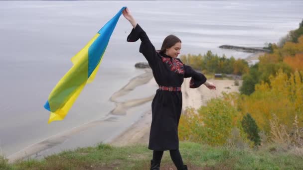 边景笑容满面的快乐女人穿着乌克兰族绣花连衣裙站在秋江畔 美丽而自信的女士在户外享受秋日 — 图库视频影像