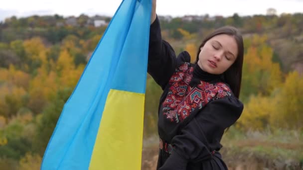 秋天阴天 年轻美丽的女子举着飘扬的乌克兰国旗站在室外 有自信的女士穿着传统服饰 有国家标志的画像 慢动作 — 图库视频影像