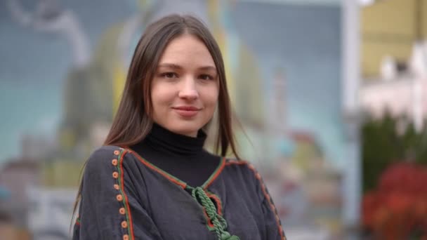 若いウクライナの幸せな女性のカメラを見て笑顔とスローモーションで周りを見回す 肖像画のブルネット自信のある女性とともに茶色の目で刺繍シャツに立って街の通り — ストック動画