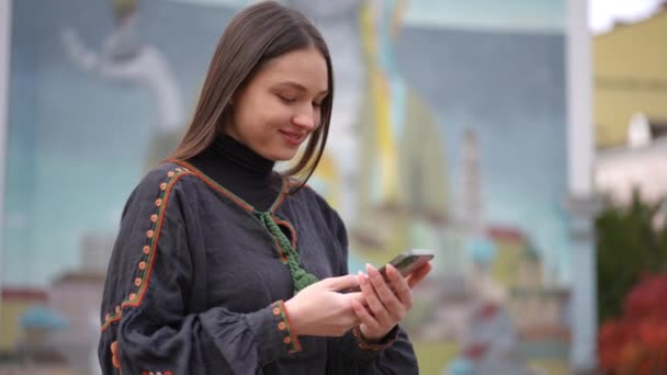 若い幸せな女性は曇りの秋の日に街の通りに立ってスマートフォンアプリでメッセージを笑顔 刺繍された黒のシャツの正のウクライナの女性の屋外 スローモーション — ストック動画