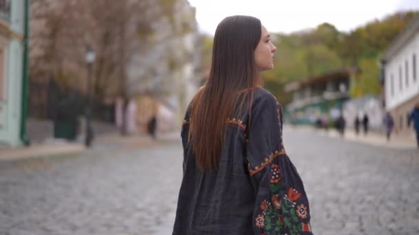 后视镜年轻瘦弱的女子沿着铺路走着 慢吞吞地环顾四周 穿着绣花衬衫在城市街道上漫步的自信美丽的乌克兰黑发女士的追踪镜头 — 图库视频影像