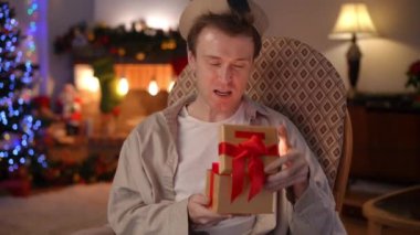 Şaşırmış yüz ifadesi olan heyecanlı LGBT adamına zum yap içinde ışık olan bir Noel hediyesi kutusu aç. Mutlu Kafkasyalı adam evinde yeni yıl hediyesiyle oturma odasında.