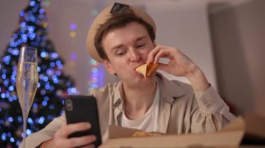 Ön görüş, milenyum LGBT adamının akıllı telefondan sosyal medyayı kaydırarak pizza yerken kafasını salladığını gösteriyor. Noel arifesinde evde genç beyaz adamın portresi.