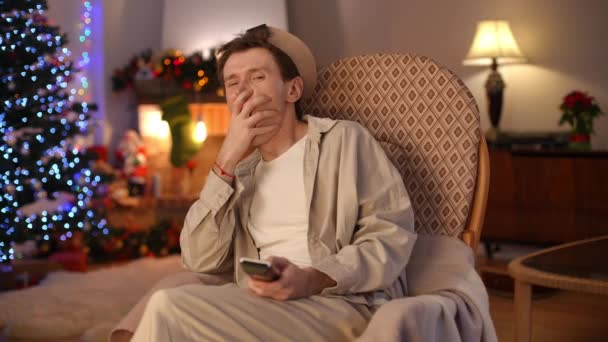 リモートコントロールでカメラを見てテレビチャンネルを切り替えるYawning興味のない男 家で一人で大晦日に休んで退屈白人Lgbtの男の肖像画 クリスマスのコンセプト — ストック動画