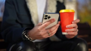 Yakın plan akıllı telefon ve kırmızı kahve fincanı tanınmayan genç adamın dövmeli ellerinde. Kafkasyalı bir adam internette mesajlaşıyor. Güneşli bir günde sonbahar parkında oturmuş sıcak bir şeyler içiyor.