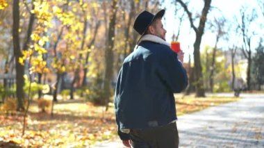Park sokağında rahatça yürüyüp kahve içen genç adamın yan görüntüsü. Kaygısız, kendine güvenen beyaz LGBT 'li adam güneşli bir sonbahar haftasonunda gezip etrafa bakıyor.