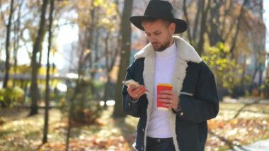 Genç, akıllı telefon uygulaması ile internetten mesajlaşan ve güz parkında gün ışığında yürüyen bir adam. Yoğunlaştırılmış yakışıklı LGBT elemanının portresi kahve fincanı ile geziniyor ve telefon uygulamasında mesajlaşıyor.