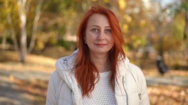 Güneşli sonbahar parkında poz veren olgun, kendine güvenen beyaz kadın portresi. Mutlu kızıl saçlı bayan dışarıda kameraya bakarak gülümsüyor.