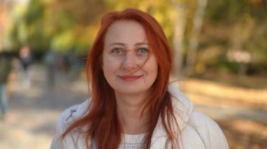 Kendinden emin olgun bir kadının güneşli bir sonbahar parkında kameraya bakarak kahve içerken çekilmiş bir fotoğrafı. Güneşli bir günde boş zamanlarında poz veren beyaz saçlı kadının Dolly portresi.