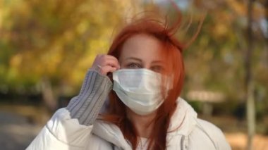 Kameraya bakan kadın gülerek iç çekerek Covid maskesini çıkarıyor. Kafkas kızıl saçlı olgun bir kadının Coronavirus salgınında sonbahar parkında poz verdiğinin ön görüntüsü. Yeni normal yaşam tarzı