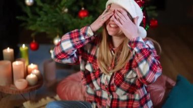 Neşeli mutlu kadın elleriyle gözlerini kapatıyor evdeki Noel ağacında oturuyor. Yeni yıl arifesinde evde sürpriz bekleyen neşeli genç bir bayanın portresi.