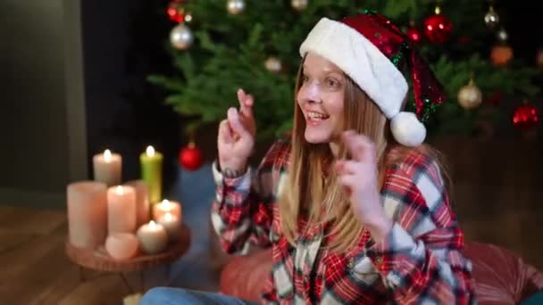 年轻快乐的女人交叉着手指说话 面带微笑地把目光移开 坐在客厅里和圣诞树在一起 快乐兴奋的白种人迷人的女士在家里的除夕夜许愿 — 图库视频影像