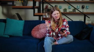 Akıllı telefon uygulamasında sosyal medyada camdan kırmızı şarap içen rahat, genç beyaz bir kadının portresi. Kaygısız büyüleyici bayanın ön görüntüsü. Evdeki hafta sonunun keyfini çıkarıyor.