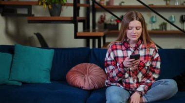 Akıllı telefonlu stresli bir kadın doğru jestle kanepede oturuyor ve sakinleşiyor. Evdeki oturma odasındaki hoşnutsuz beyaz kadının portresi.