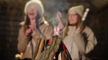 Yakın çekim şenlik ateşi, arka planda yavaş çekimde dans eden, bulanık, beyaz, neşeli genç kadınlarla Noel şarkısı söyleyerek yanıyor. Tanınamayan mutlu arkadaşlar dışarıda tatil kutlarken eğleniyorlar.