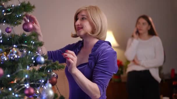满意的性别流线型女人装饰新年树 模糊的女性朋友在电话背景下交谈 积极轻松的白人非二元人在家里庆祝节日 — 图库视频影像