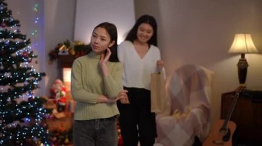 Düşünceli genç, güzel Asyalı bir kadın Noel arifesinde oturma odasında arka planda bir arkadaş olarak duruyor. Elinde hediyeyle alışveriş çantasından geçiyor. Heyecanlı bir bayan yeni yıl hediyesi için teşekkür ediyor.