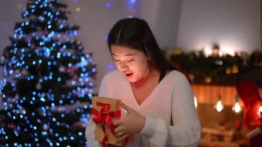 Heyecanlı Asyalı kadının portresi içinde ışık olan hediye kutusunu açıyor ve başka tarafa bakıyor. Mutlu sürpriz genç bayan Noel hediyesiyle yılbaşı arifesinde evde.