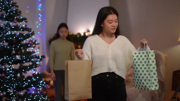 一个兴奋的亚洲迷人女人的画像 她头戴购物袋 模糊的朋友微笑着 双手叉腰站在壁炉边 背靠着圣诞树 快乐的女士在室内摆姿势 — 图库视频影像