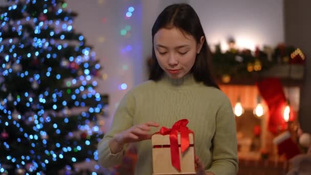 在圣诞前夕 一位身材苗条迷人的亚洲年轻女子站在客厅里 带着惊讶的表情打开礼品盒 千禧年女子的画像 带着淡淡的微笑欣赏着礼物 — 图库视频影像
