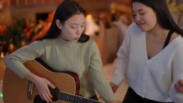 新年前夕 两个快乐的亚洲女人坐在室内弹奏吉他手的高角肖像 有才华 有自信 快乐的朋友们 在圣诞快乐地享受着自己的业余爱好 — 图库视频影像