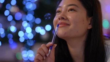 Noel arifesinde evde Noel Baba 'ya mektup yazan gülümseyen Asyalı kadının yakın plan portresi. Mutlu, kendinden emin, pozitif düşünceli bir kadının Noel ışıklarında evde kalemle yattığını gösteren bir vesikalık.