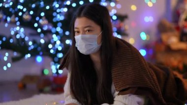 Coronavirus 'lu genç Asyalı kadın maskeyle öksürüyor ve içeride Noel ışığı altında yattığını düşünüyor. Yeni yıl arifesinde karantinada yalnız üzgün bir kadının portresi.